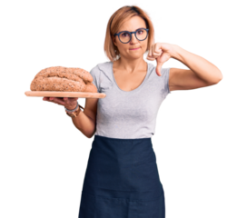 Young blonde woman holding wholemeal bread with angry face, negative sign showing dislike with thumbs down, rejection concept