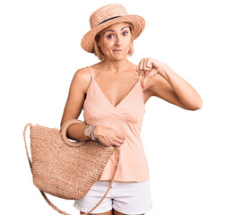 Young blonde woman wearing summer hat holding bag with angry face, negative sign showing dislike...