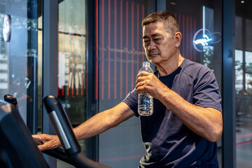 Asian elderly old man in the 70s or 80s drinking water after doing exercise in the gym - a health...