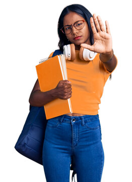 Young indian girl holding student backpack and books with open hand doing stop sign with serious and confident expression, defense gesture
