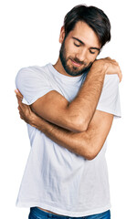 Hispanic man with blue eyes wearing casual white t shirt hugging oneself happy and positive, smiling confident. self love and self care