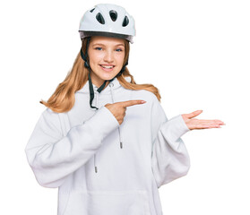 Beautiful young caucasian girl wearing bike helmet amazed and smiling to the camera while presenting with hand and pointing with finger.