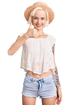 Young blonde woman with tattoo wearing summer hat doing happy thumbs up gesture with hand. approving expression looking at the camera showing success.