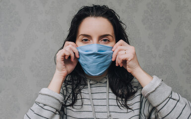 A young woman corrects the blue protective medical mask-respirator on gray background. The concept...