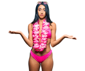Beautiful hispanic woman wearing bikini and hawaiian lei clueless and confused expression with arms and hands raised. doubt concept.