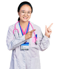 Young chinese woman wearing doctor uniform and stethoscope smiling and looking at the camera pointing with two hands and fingers to the side.