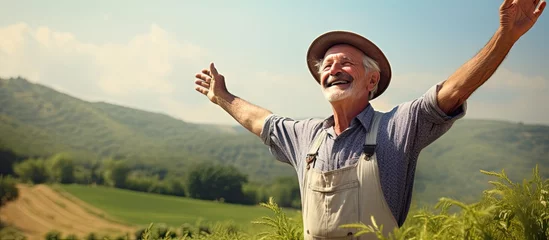 Fotobehang Joyful elderly man farmer stands on farmland, arms raised in happiness. © AkuAku