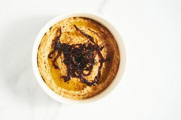 Hummus with caramelized onions in white bowl on light background