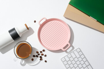 A cup of coffee, a pink ceramic tray, a keyboard, coffee beans and a thermos are displayed together on a white background. Caffeine is a natural stimulant found in coffee.