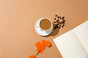 A cup of milky coffee, coffee beans, a notebook and a branch of autumn leaves are displayed on a brown background. Coffee beans contain many powerful antioxidants.