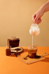 The hand is holding a glass of milk pouring into a cup of coffee, surrounded by coffee beans,...