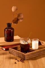 Autumn leaves are inserted in a brown vase, on a wooden tray there is a cup of coffee and a cup of milk, coffee beans scattered on the wooden table. Space for coffee advertising.
