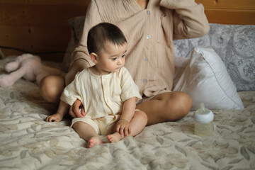 Fototapeta premium A baby sits thoughtfully on a bed, the gentle presence of a mother nearby. It s a calm moment in the rhythm of family life.