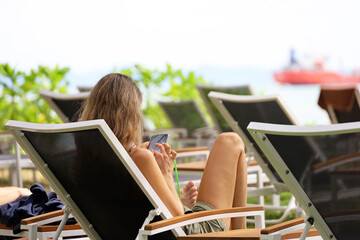 Girl sitting with smartphone in hands in deck chair on sea background. Online communication on...
