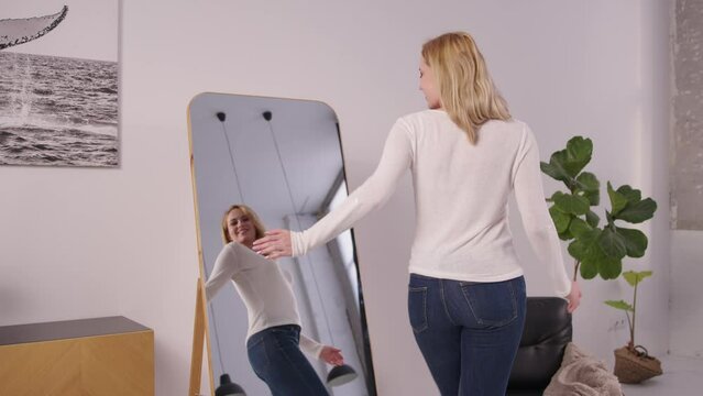 Portrait of joyful charming mature woman in trendy clothes looking at reflection in mirror, enjoying appearance and shapes, slapping on buttocks', expressing cheerful mood and excitement.