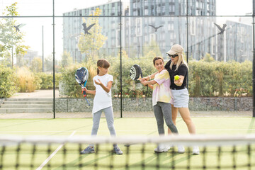 Young tennis trainer in activewear holding racket held by one of two cute girls while consulting...