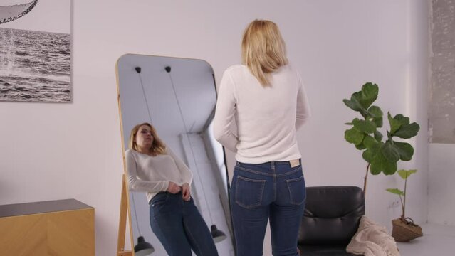 Irritated pretty mature women with weight problems trying to button up tight jeans, expressing despair and disappointment while standing in front of mirror indoors