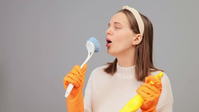 Domestic chores and cleaning service. Glad young Caucasian housewife holding cleaning brush and bottle of detergent doing housework isolated over gray background having fun singing with brush
