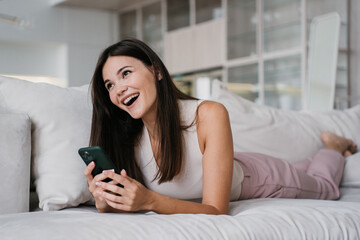 Brunette cheerful girl laying on couch at home using phone laughing watching funny videos on social media feed. Weekend, female relaxing indoors.