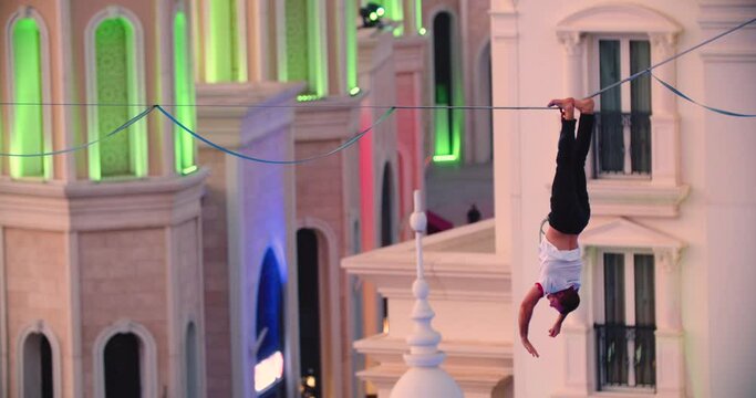 Slackliner is hanging upside down on the slackline between buildings, 4k