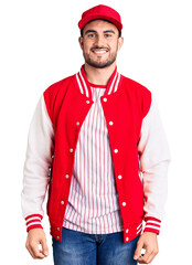 Young handsome man wearing baseball jacket and cap looking positive and happy standing and smiling with a confident smile showing teeth