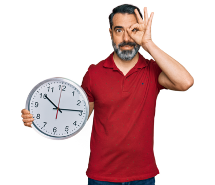 Middle aged man with beard holding big clock smiling happy doing ok sign with hand on eye looking through fingers