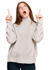Young read head woman wearing casual winter sweater amazed and surprised looking up and pointing with fingers and raised arms.