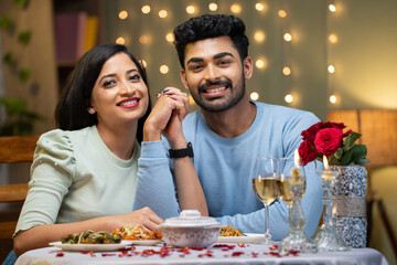 Happy smiling couple holding hands by looking at camera during candle light dinner at home - concept of valentine's day, love and relationship.