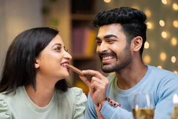 Happy young man giving chocolate byte to girlfriend during candle light dinner at home - concept of wedding anniversary, romantic moment and affection