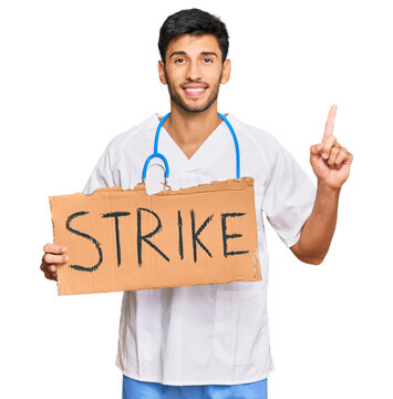 Young handsome man wearing doctor uniform holding strike banner cardboard surprised with an idea or question pointing finger with happy face, number one