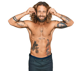 Handsome man with beard and long hair standing shirtless showing tattoos smiling pulling ears with...
