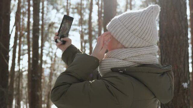 Young woman admires and preens herself at smartphone camera in sunny winter forest, handheld shot. Lady smiles and takes selfie on frosty day. Single female creates photo for dating app.