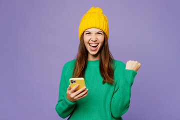 Young woman she wear green sweater yellow hat casual clothes hold in hand use mobile cell phone do winner gesture isolated on plain pastel light purple background studio portrait. Lifestyle concept.