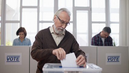 A senior man is throwing a voting ballot into the box at the polling station, actively...