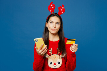 Merry minded young Latin woman wear red Christmas sweater decorative deer horns on head posing hold...