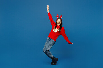 Full body merry young Latin woman wear red Christmas sweater fun decorative deer horns on head posing stand on toes lean back dance isolated on plain blue background. Happy New Year holiday concept.