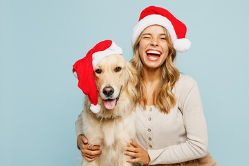 Young joyful owner woman wears casual clothes Santa hat hug cuddle embrace best friend pet retriever dog wink isolated on plain pastel blue background studio. New Year Christmas celebration concept.