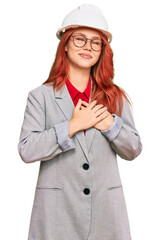 Young redhead woman wearing architect hardhat smiling with hands on chest with closed eyes and grateful gesture on face. health concept.