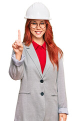 Young redhead woman wearing architect hardhat showing and pointing up with finger number one while smiling confident and happy.