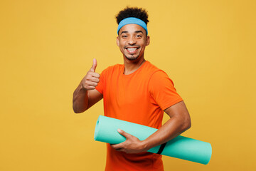 Young fitness trainer instructor sporty man sportsman wear orange t-shirt hold in hand yoga mat show thumb up training in home gym isolated on plain yellow background. Workout sport fit abs concept.