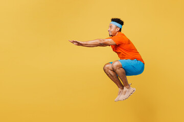 Full body side profile view young fitness trainer sporty man sportsman wear orange t-shirt jump high do squat spend time in home gym isolated on plain yellow background. Workout sport fit abs concept.