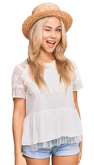 Young blonde girl wearing summer hat winking looking at the camera with sexy expression, cheerful...