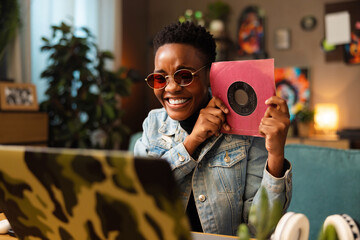 Portrait of dark skinned young woman in pink sunglasses smiling holding cd in hands showing at...