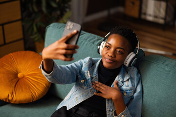 Happy delighted afro american woman with short haircut wearing new modern headphones holding cell...