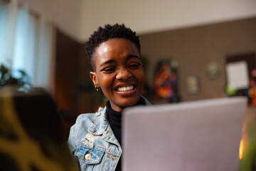 Attractive cute afro american dark skinned woman with short hair smiling holding papers in hands observing working studying in front of computer laptop.
