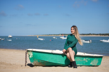 Fototapeta premium Young, pretty, blonde, green-eyed young woman in a green dress, sitting in a boat, looking at the camera, relaxed and calm, on the beach, with the sea in the background. Concept vacation, sun, travel