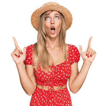 Beautiful caucasian woman wearing summer hat amazed and surprised looking up and pointing with fingers and raised arms.