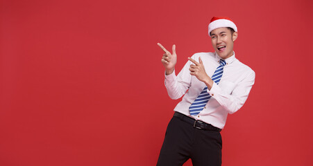Happy asian business man wearing santa hat pointing finger isolated on red copy space background....