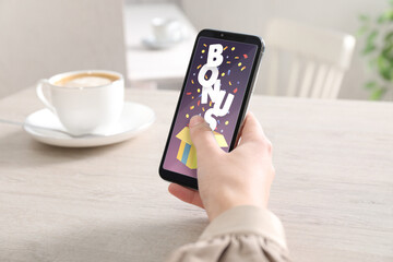 Bonus gaining. Woman holding smartphone at white wooden table, closeup. Illustration of open gift box, word and confetti on device screen