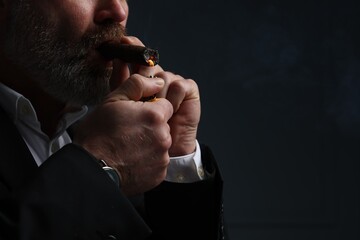 Bearded man lighting cigar on dark grey background, closeup. Space for text